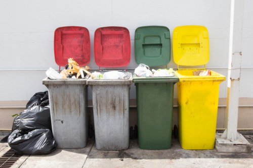A row of four large plastic wheelie bins positioned on a paved outdoor surface against a plain light-colored wall. From left to right, the first bin is grey with an open red lid and topped with a pile of mixed waste including paper and plastic packaging. Next, the second grey bin has a closed red lid with some rubbish piled on top, such as cardboard and plastic waste. The third bin is green with a closed green lid, also topped with some loose rubbish. The fourth bin is yellow, with a closed yellow lid, and has bits of packaging waste spilling over the top. To the left of the bins, a tightly packed black trash bag leans against the grey bin. The environment appears to be an outdoor storage area, possibly part of a commercial property or waste disposal zone, fitting within waste removal services for the Abbey Wood area. This arrangement of bins and waste demonstrates typical rubbish collection and disposal setups managed by professionals like Commercial Waste Removal Abbey Wood.