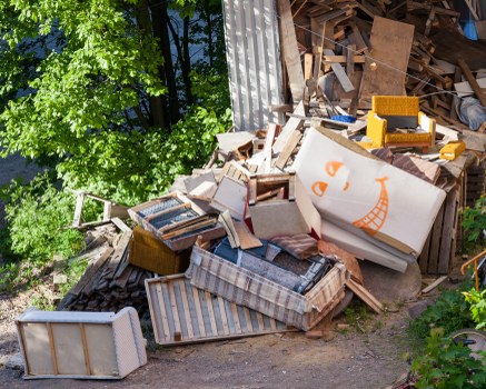 A large pile of discarded household furniture and wooden debris outdoors next to a corrugated metal structure. The waste includes an old striped armchair, a wooden cabinet, broken wooden planks, and various unassembled furniture pieces, some with visible upholstery or wood finishes. Bright sunlight illuminates the scene, with green foliage and trees visible on the left side, indicating the waste is located on a driveway or outdoor space near a residential property. The clutter appears to be in the process of disposal, suitable for rubbish removal services offered by Commercial Waste Removal Abbey Wood, serving the local area near Abbey Wood. The materials are predominantly wood and upholstery, with a mix of textures from smooth painted surfaces and rough cut edges, arranged in a scattered manner, suggesting collection or clearance activities in progress.