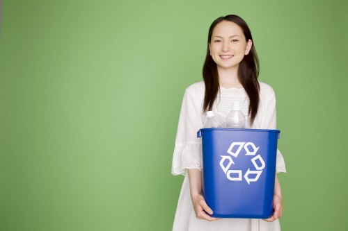 A young woman with long dark hair and a friendly expression, wearing a white lace dress, is standing against a solid light green background. She is holding a blue plastic recycling bin with the universal recycling symbol printed in white on the front, positioned centrally in her hands. The bin appears to be empty or lightly filled, and she is gripping it with both hands at the sides. The setting is free of additional objects or environmental clutter, emphasizing the bin's design and her engagement with recycling. The scene is well-lit, with soft, even lighting that highlights the bright colours and clear textures of the bin and her attire. This image visually supports waste management and rubbish disposal themes relevant to commercial waste removal services in Abbey Wood, illustrating a focus on environmentally conscious practices without depicting specific rubbish or waste materials.