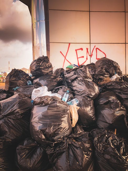 A large pile of black garbage bags, filled with waste, stacked against a tiled wall in an urban outdoor setting. Several of the bags are tied at the top, with some appearing bulging and heavy, and a f